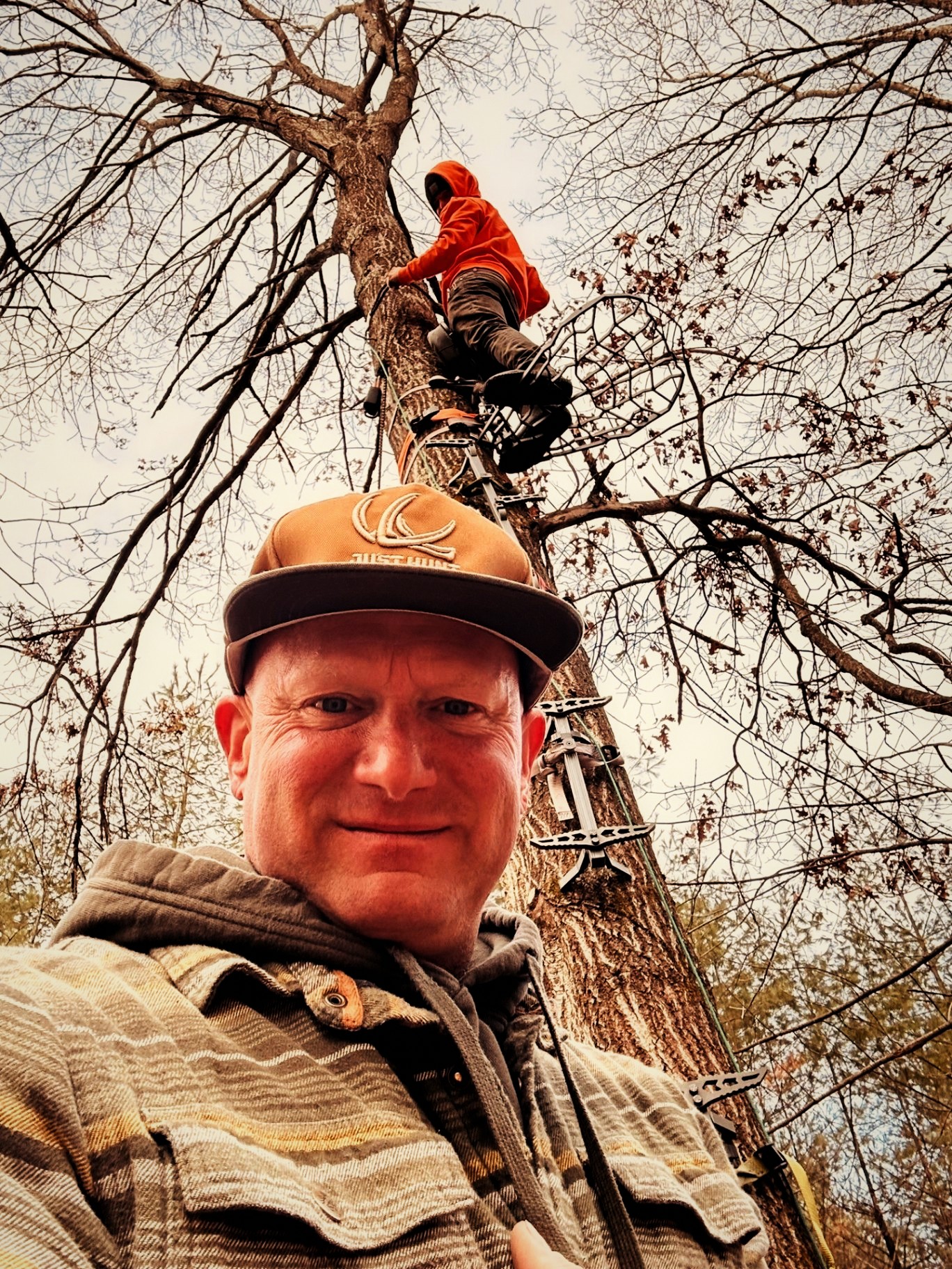 Hunter grinning below a climbing partner setting a treestand in bare spring timber