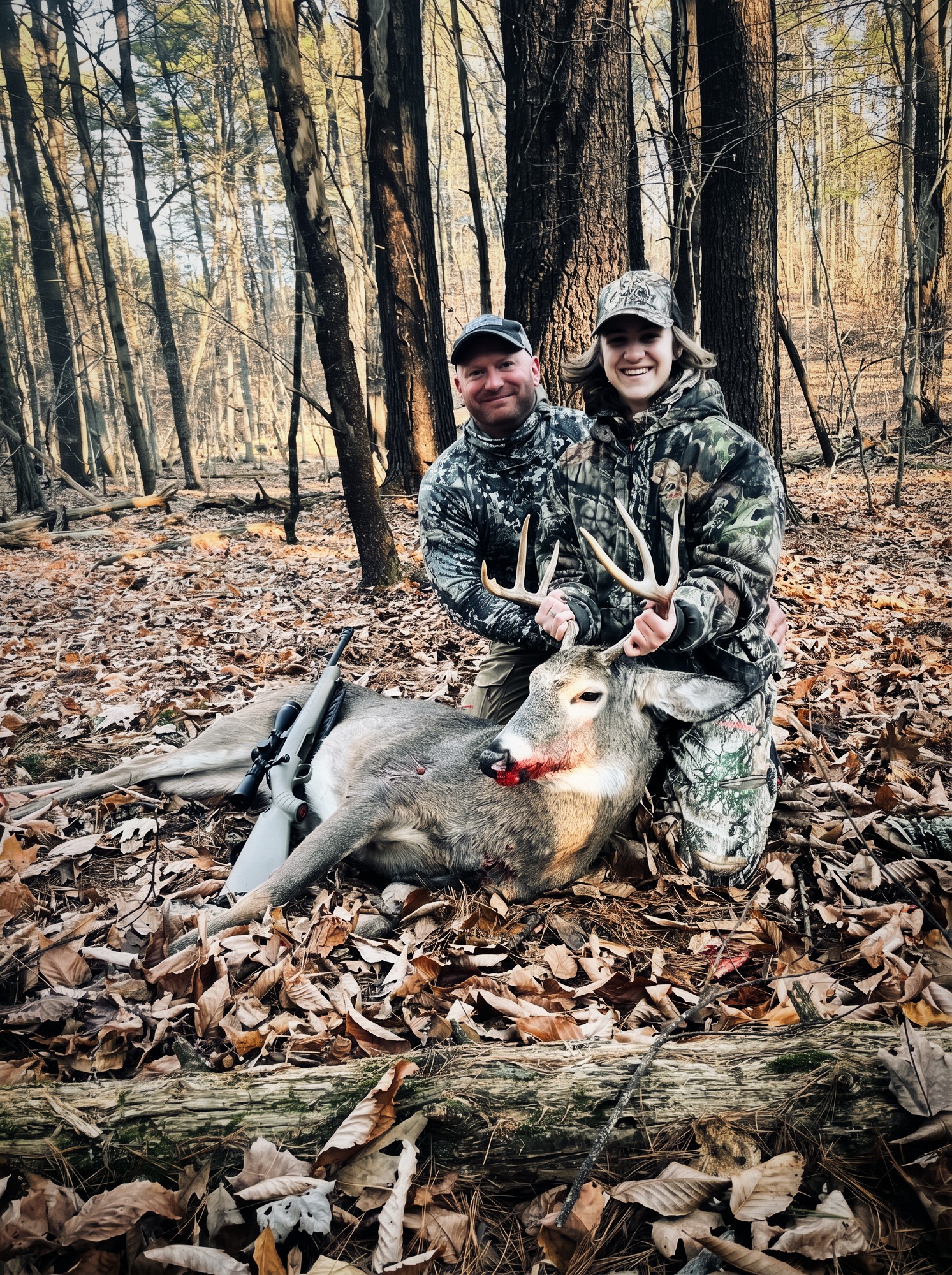 Two hunters kneeling behind a harvested whitetail buck in hardwood timber