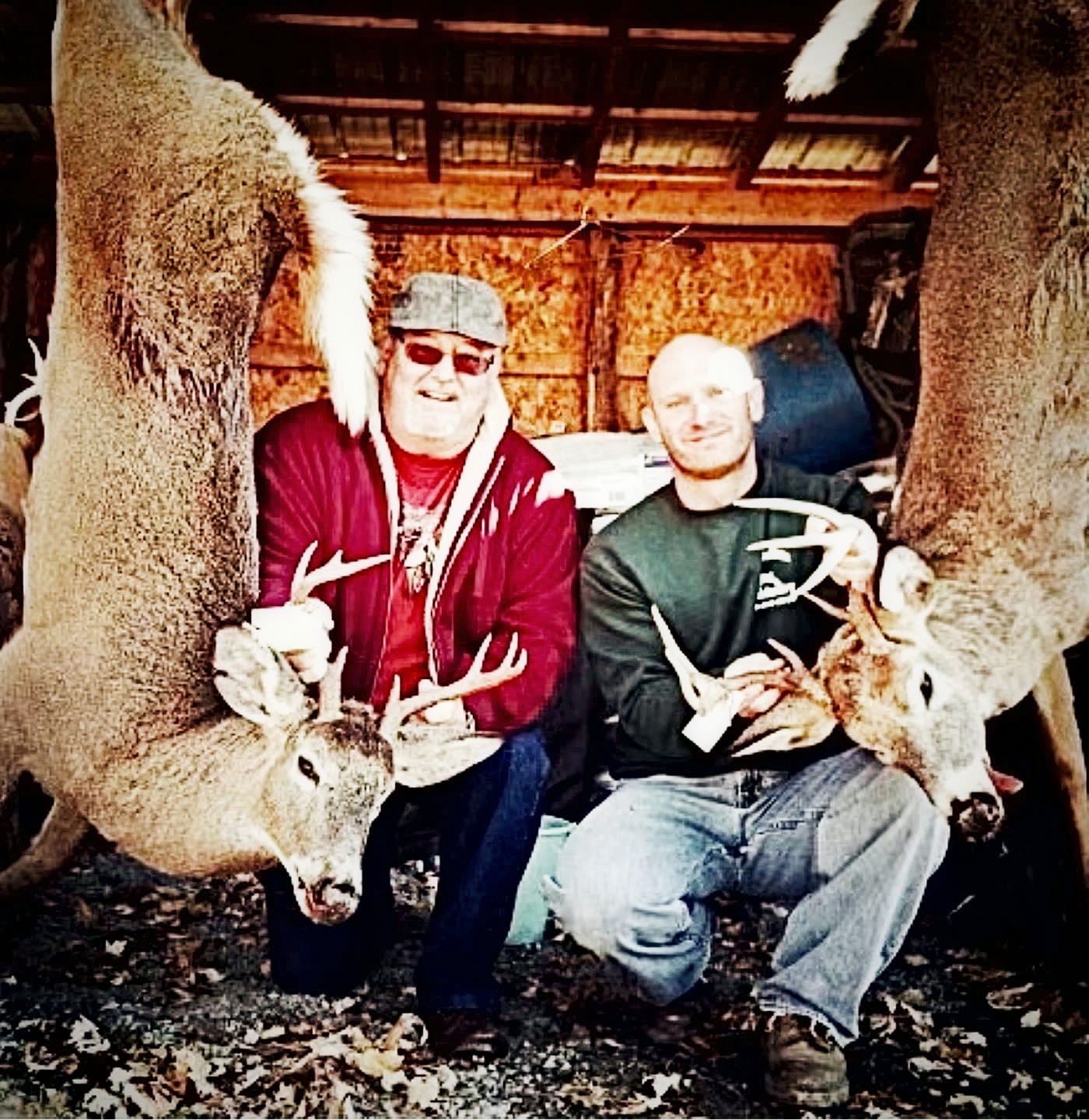 Two hunters kneeling between hanging whitetail bucks in an old barn